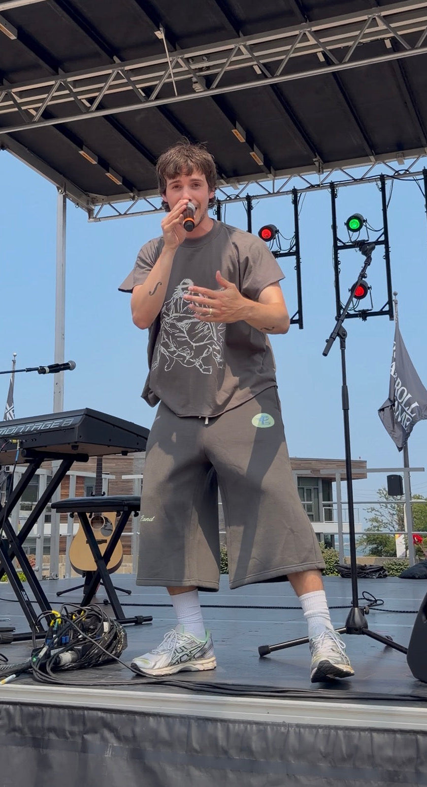 Conor Michael Smith performing live on an outdoor stage, wearing a gray graphic t-shirt, matching gray shorts, white socks, and sneakers, holding a microphone.