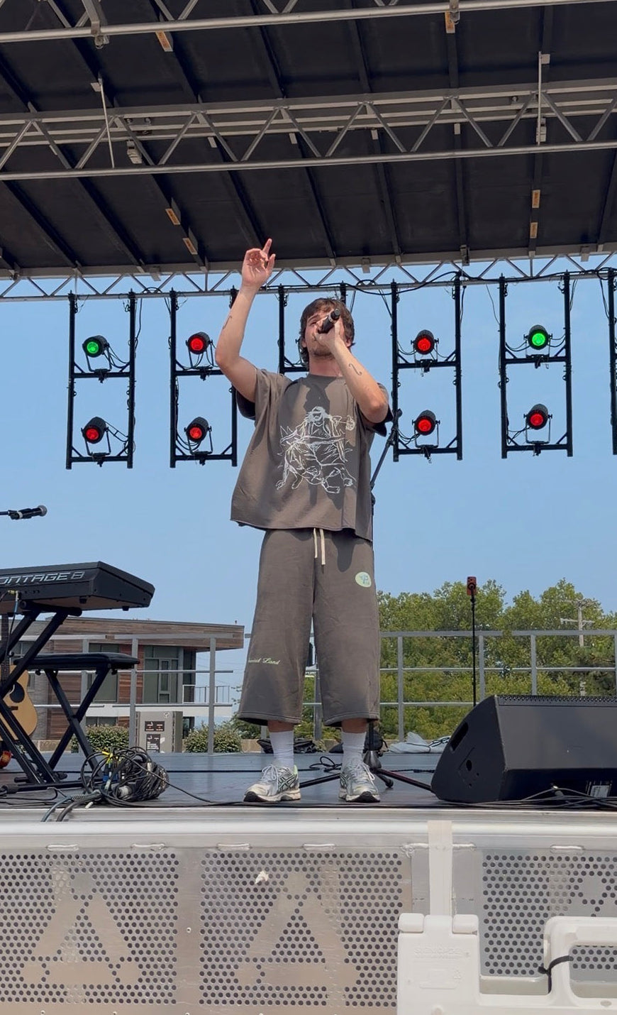 A person stands on an outdoor stage under a metal canopy, holding a microphone and raising one arm. They are wearing a loose gray graphic T-shirt, matching gray sweatpants, white socks, and sneakers.